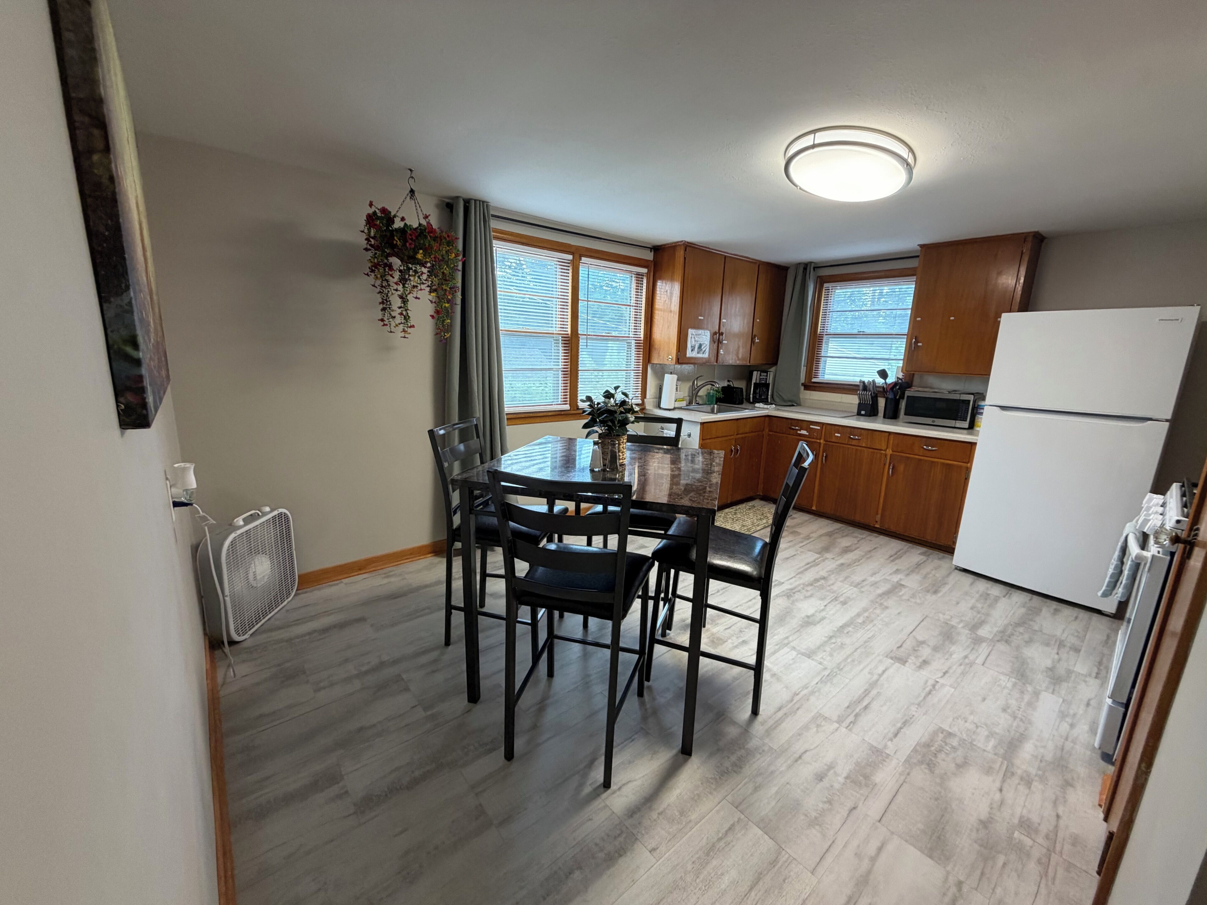 A compact kitchen with a dining table, wooden cabinets, a refrigerator, and a window with blinds.