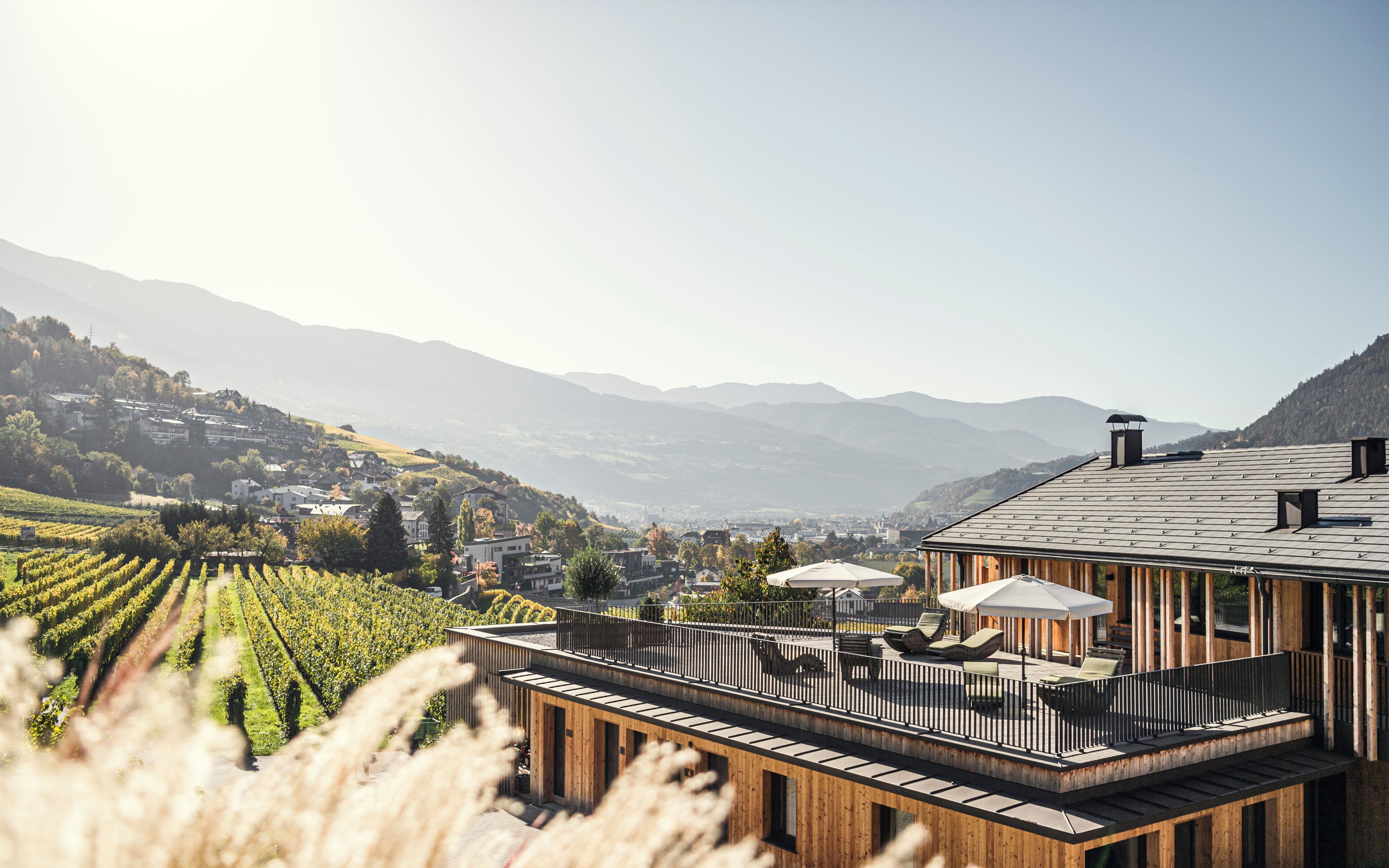 Ein modernes Gebäude mit einer Holzterrasse und Sitzgelegenheiten im Freien, mit Blick auf einen Weinberg und entfernte Berge.