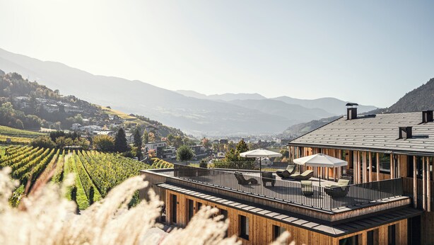 Un edificio moderno con terraza de madera y asientos exteriores, con vistas a un viñedo y montañas distantes.