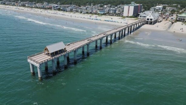 Eine Pier, die sich ins Wasser erstreckt, mit einem Gebäude am Ende, ein Strand mit Menschen und Häuser im Hintergrund.