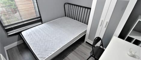 A small, single-bed room with a metal headboard, a white desk, and a chair. There is a window with blinds, a wardrobe with mirrored doors, and a radiator on the floor.