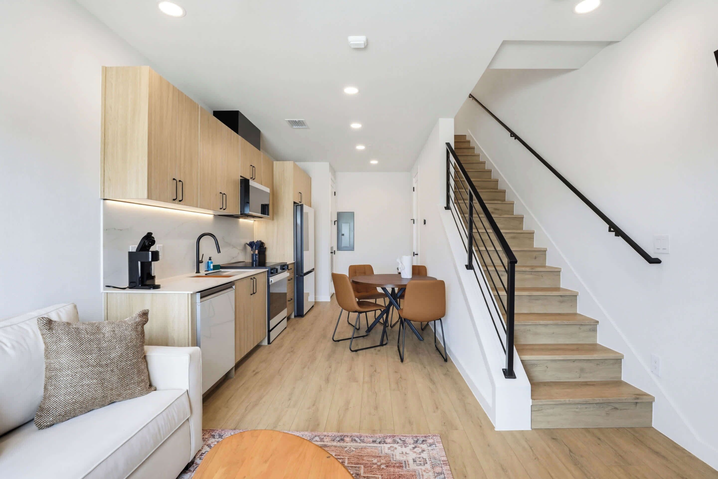 Una cocina moderna con armarios de madera, un comedor con mesa y sillas, una escalera y una pared blanca.