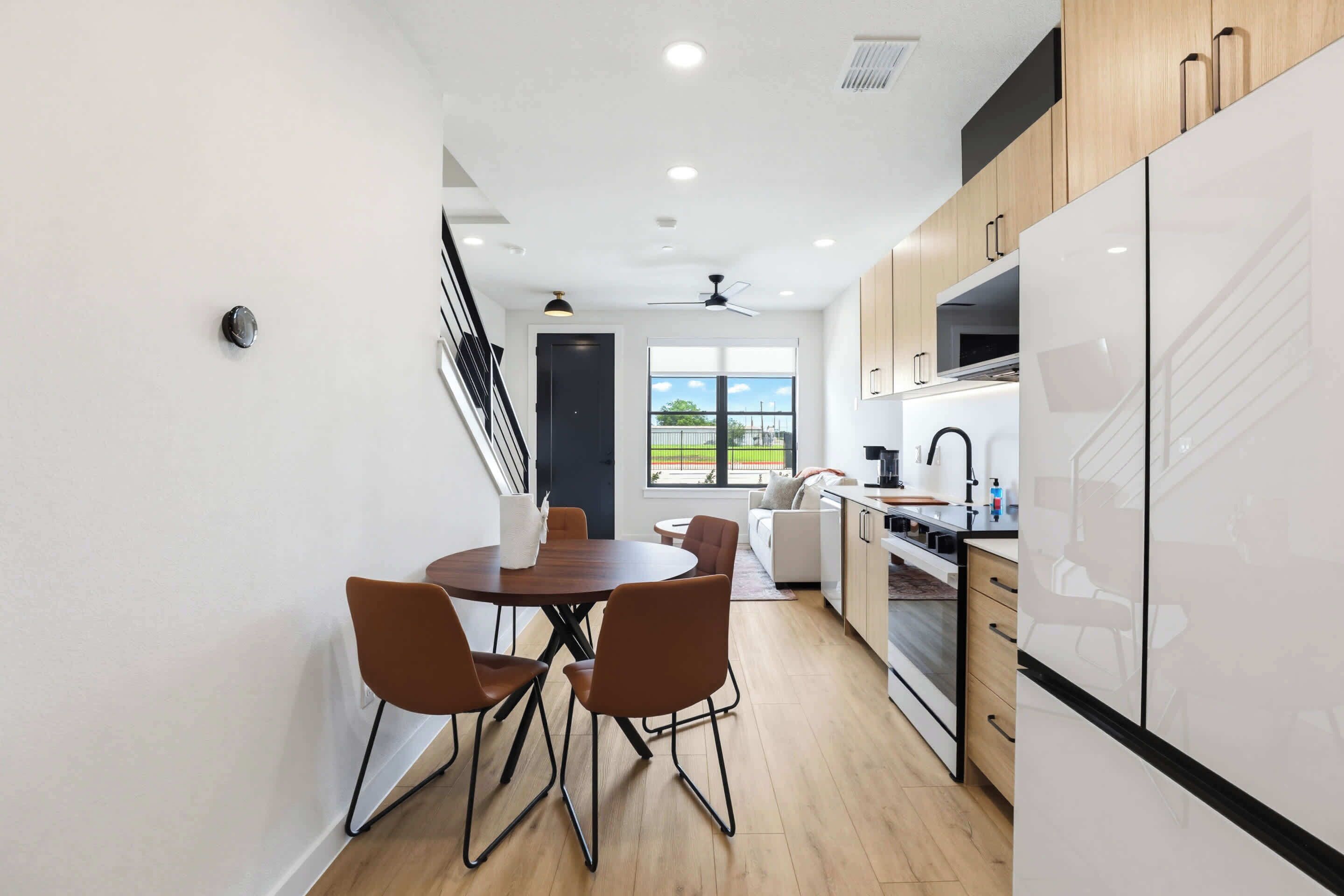 A modern kitchen with white cabinets, a dining area with brown chairs, and a view through the window.