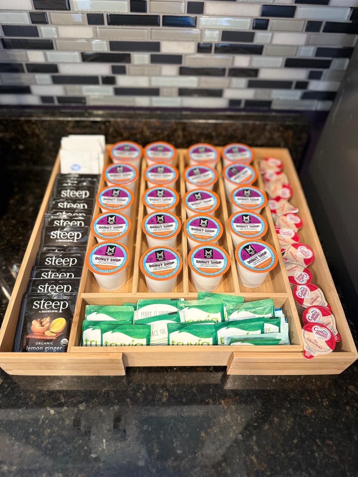 Een houten dienblad met diverse verpakte producten, waaronder dozen met de aanduiding 'steep' en 'biologische citroen-gember', en bakjes met 'Donut Shop' branding.