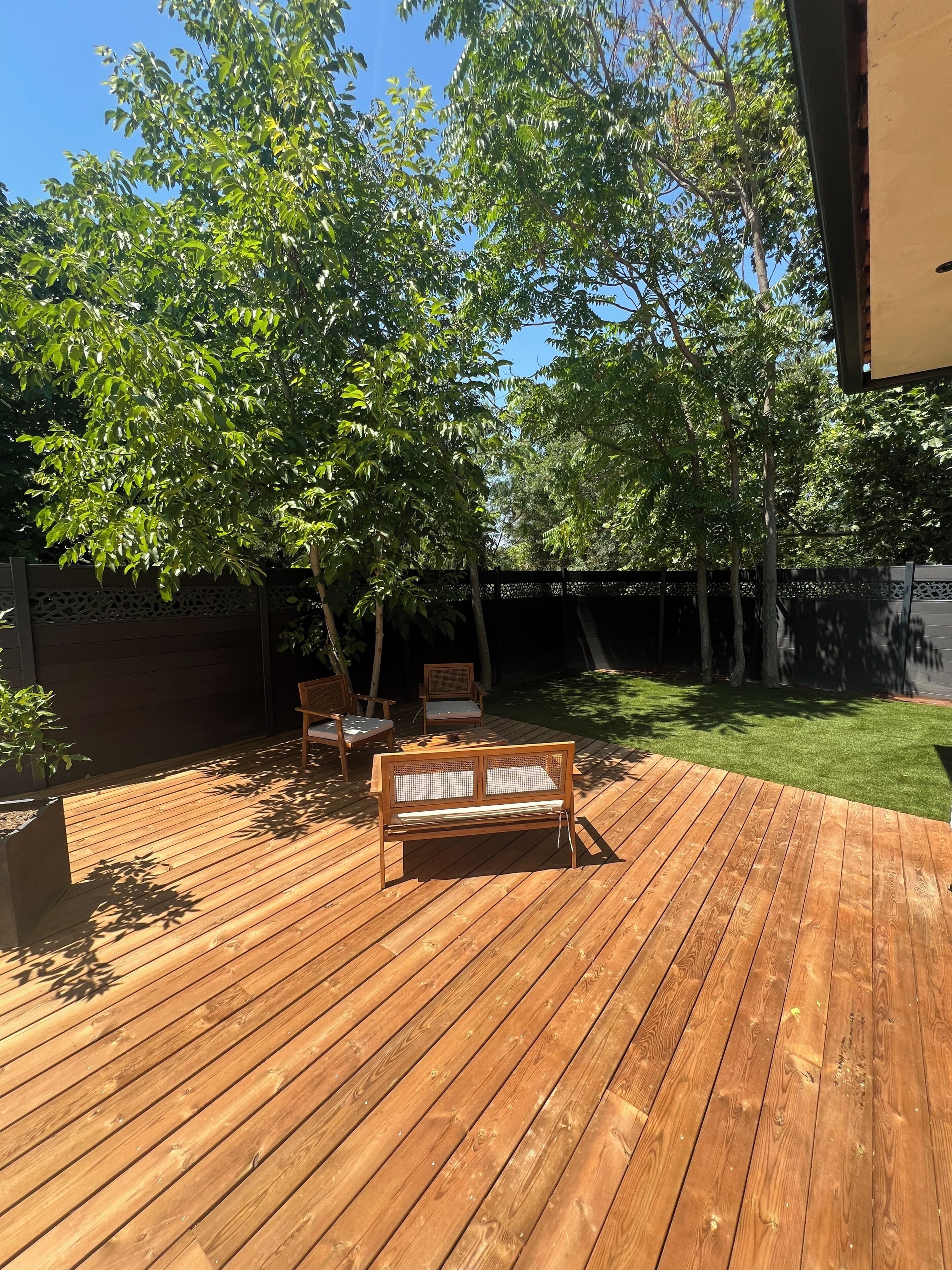 Een houten terras met een bank en stoelen, omgeven door bomen en een verzorgd gazon.