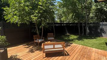 Een houten terras met een bank en stoelen, omgeven door bomen en een verzorgd gazon.