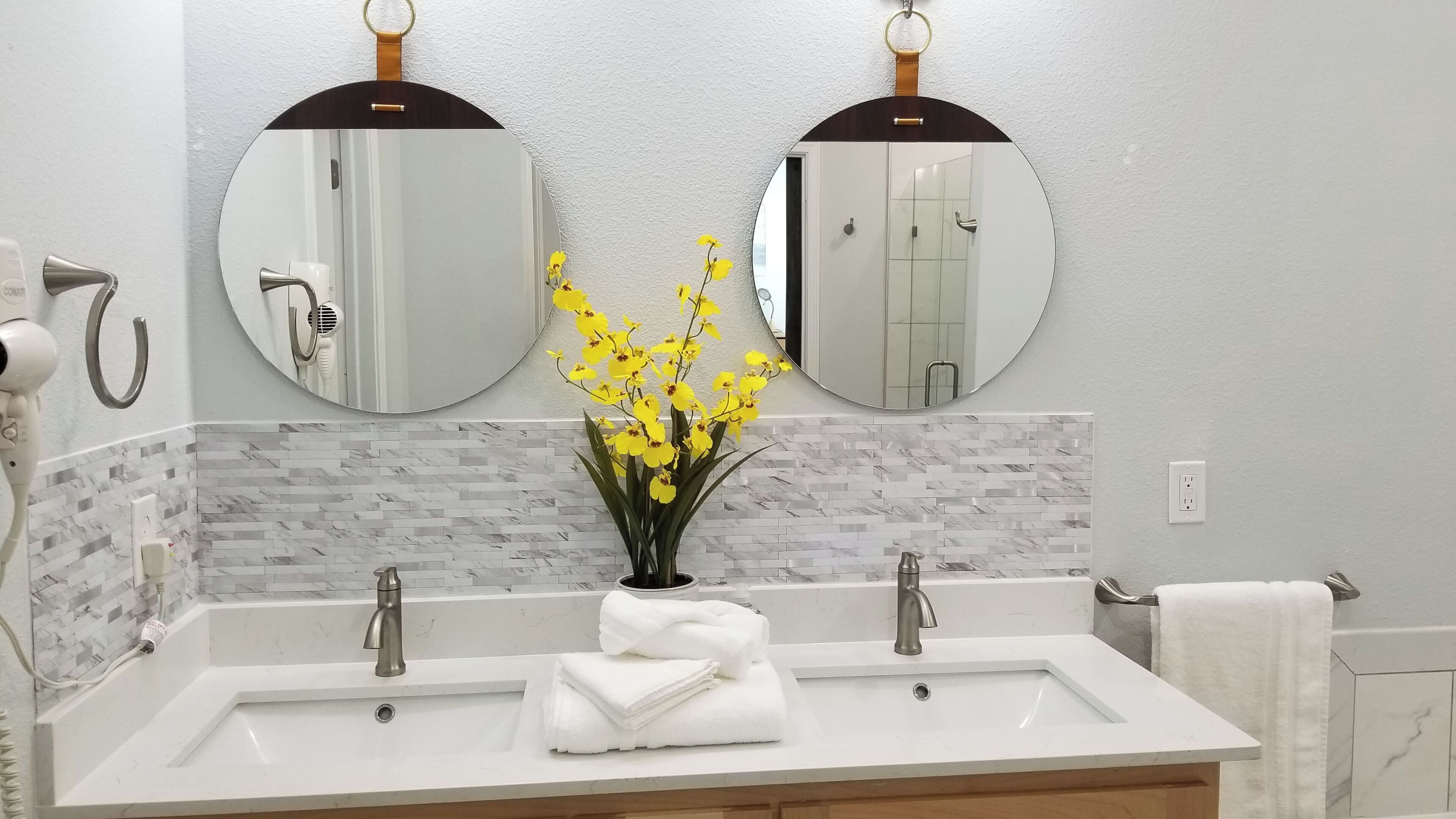 Une salle de bain avec deux lavabos, un miroir, un porte-serviettes et un vase rempli de fleurs jaunes.