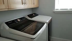 A front-loading white washer and dryer unit with a control panel, situated in a room with light gray walls and wooden cabinets above.