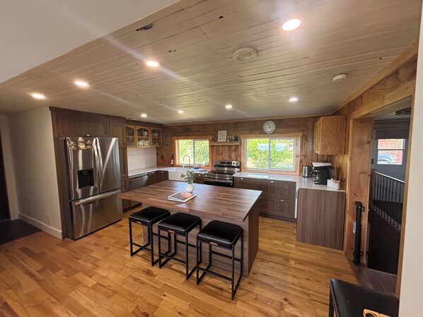 A modern kitchen with wooden cabinets, stainless steel appliances, a large island with stools, and a view of the outdoors through a window.