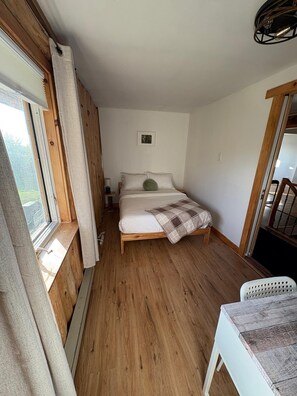 Une chambre avec un plancher en bois, un lit avec une literie blanche, une fenêtre avec des rideaux et une petite table avec une chaise.