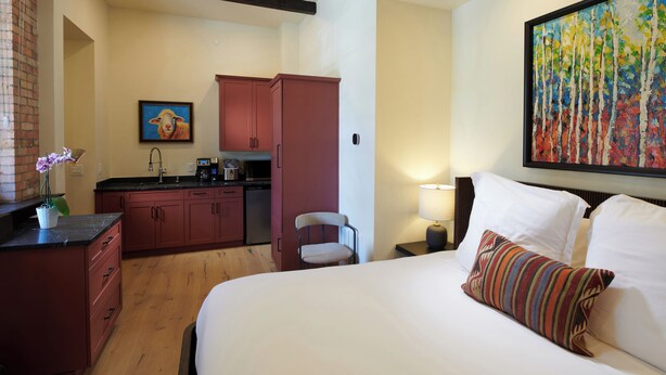 A hotel room with a bed, a desk, a chair, a small kitchenette, and a painting on the wall.