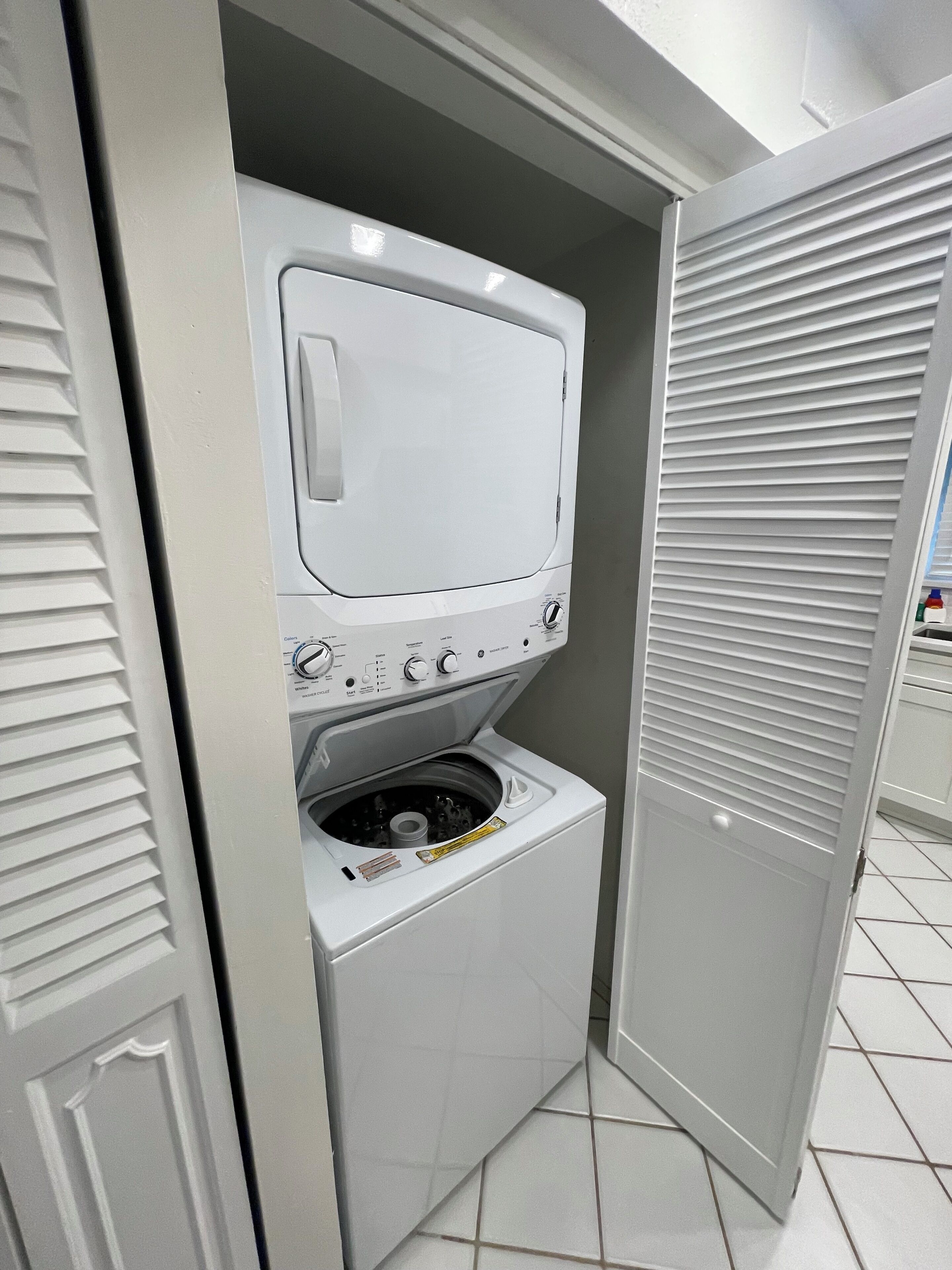 Een kleine wasruimte met een wasmachine en droger, afgesloten door een grijze deur met een verstelbaar ventilatierooster.