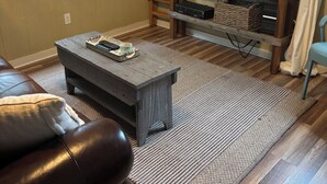 Una sala de estar con piso de madera, un televisor sobre un mueble de madera, una mesa de centro, un sofá de cuero y una ventana con vista a la vegetación.