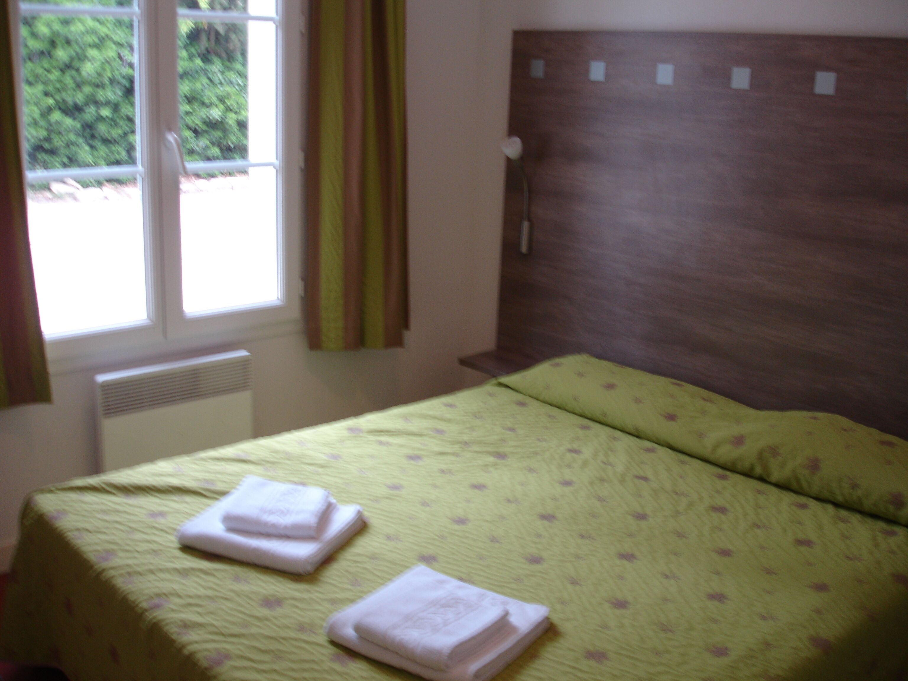 Une chambre à coucher comprenant un lit, une fenêtre, des rideaux, un radiateur et un luminaire fixé au mur.