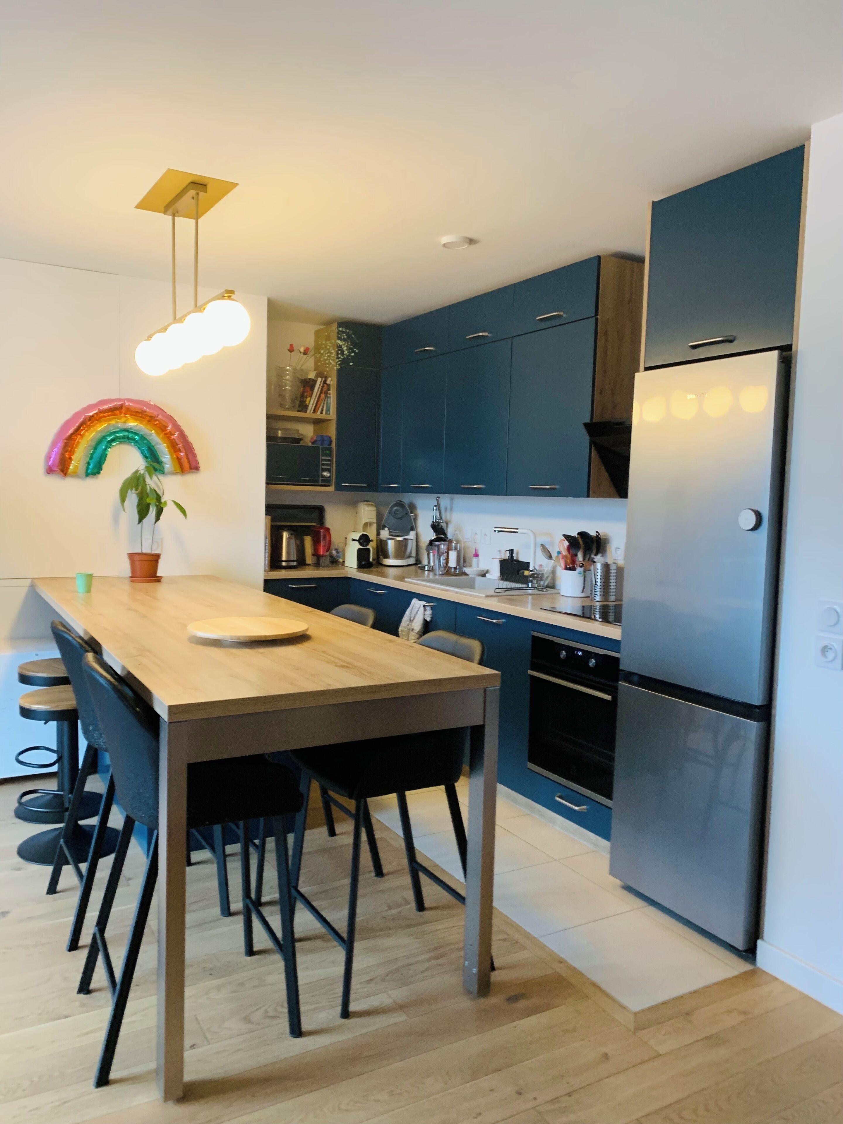 Eine moderne Küche mit dunkelblauen Schränken, einem Edelstahlkühlschrank, einem Holzespeisebereich mit Stühlen und einer farbenfrohen Lichtdekoration.