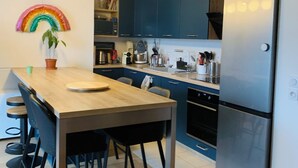 Een moderne keuken met donkerblauwe kastjes, een roestvrijstalen koelkast, een houten eettafel met stoelen en een lichtdecoratie in regenboogkleuren.