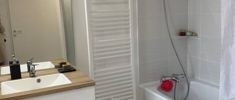 Une salle de bain moderne avec un meuble-lavabo blanc, un grand miroir, une douche équipée d’un pommeau de douche à main et un porte-serviettes blanc.