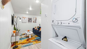 Sala de estar moderna com sofá, mesa de centro e objetos decorativos. Uma máquina de lavar roupa é visível ao fundo.