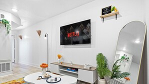 Uma sala de estar moderna com televisão de tela plana, um sofá branco, uma mesa de centro, um espelho e uma prateleira pequena com objetos decorativos.