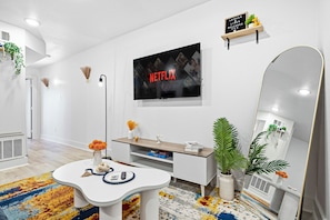 Una sala de estar moderna con un televisor de pantalla plana, un sofá blanco, una mesa de centro, un espejo y un pequeño estante con adornos.
