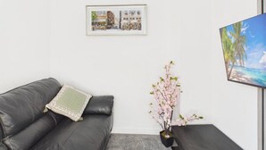 A room with a dark leather sofa, a small table with a vase of pink flowers, a framed picture on the wall, and a large framed picture on the right.