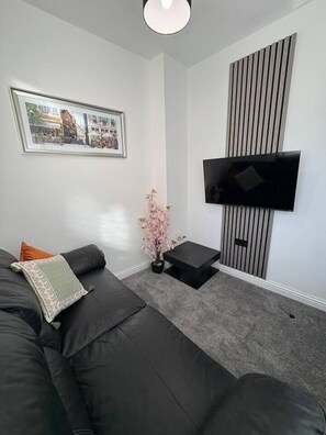 A modern living room with a black leather sofa, a flat-screen TV mounted on the wall, a small side table, and a framed picture on the wall.