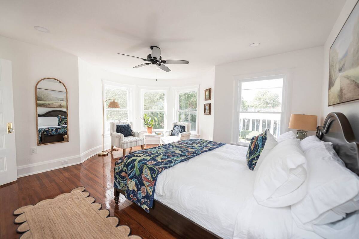 A bedroom with a large bed, a ceiling fan, a window with a view, a chair, and a rug.