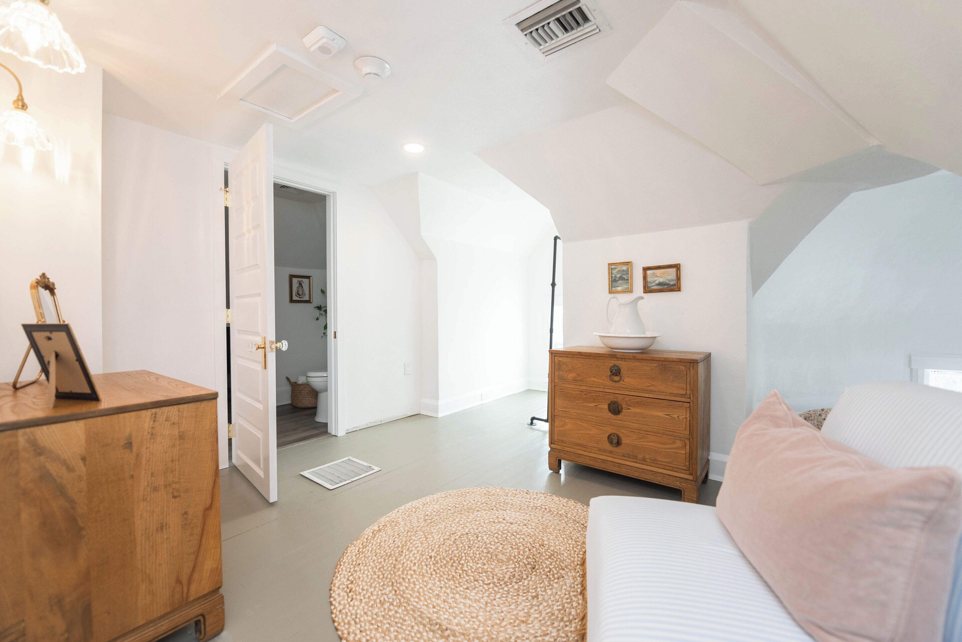 A bedroom with a bed, a wooden dresser, a round ottoman, a nightstand, and a wall-mounted lamp.