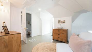 Een slaapkamer met een bed, een houten dressoir, een rond poefje, een nachtkastje en een wandlamp.
