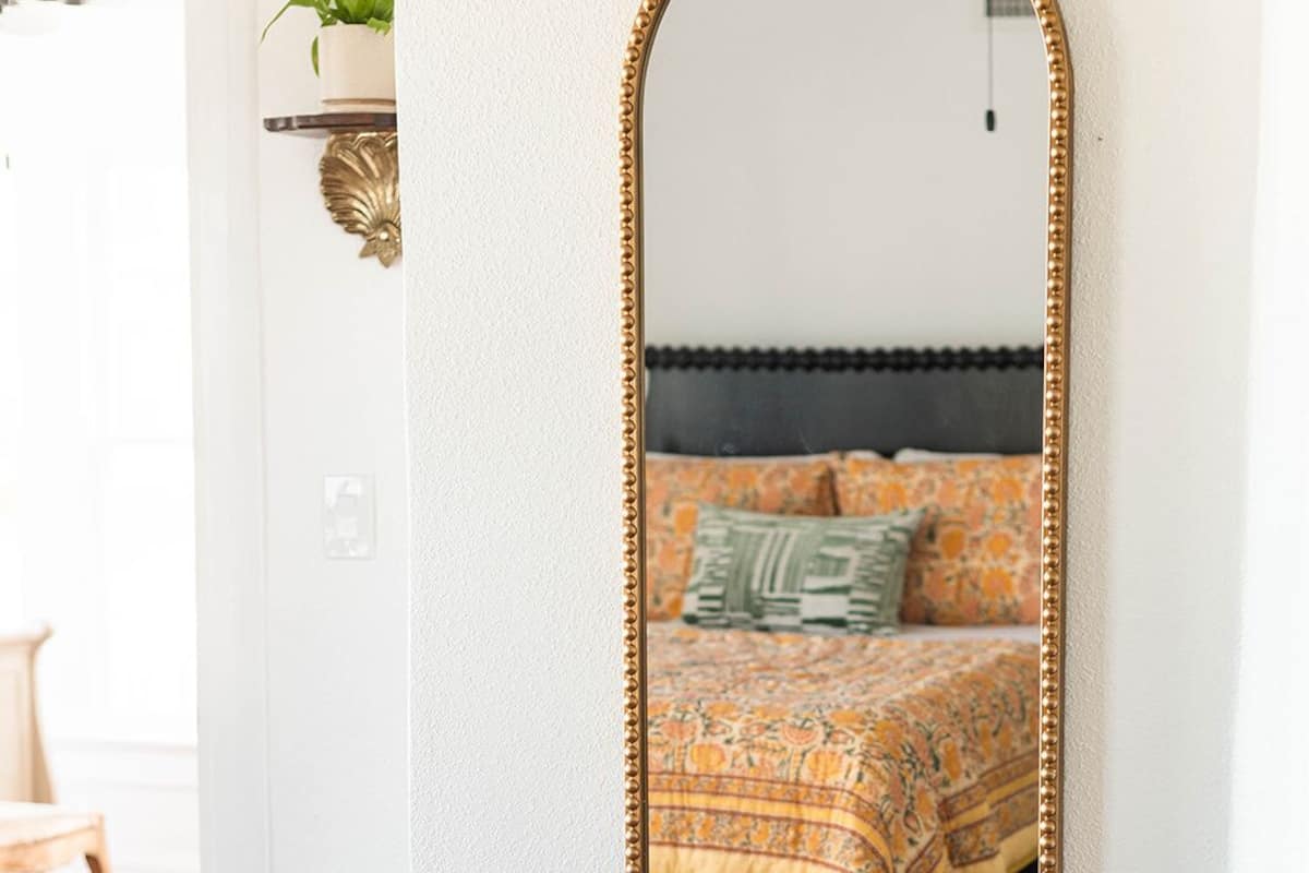 A bedroom with a large mirror, a bedside table, a bed with patterned bedding, and a green plant on a shelf.