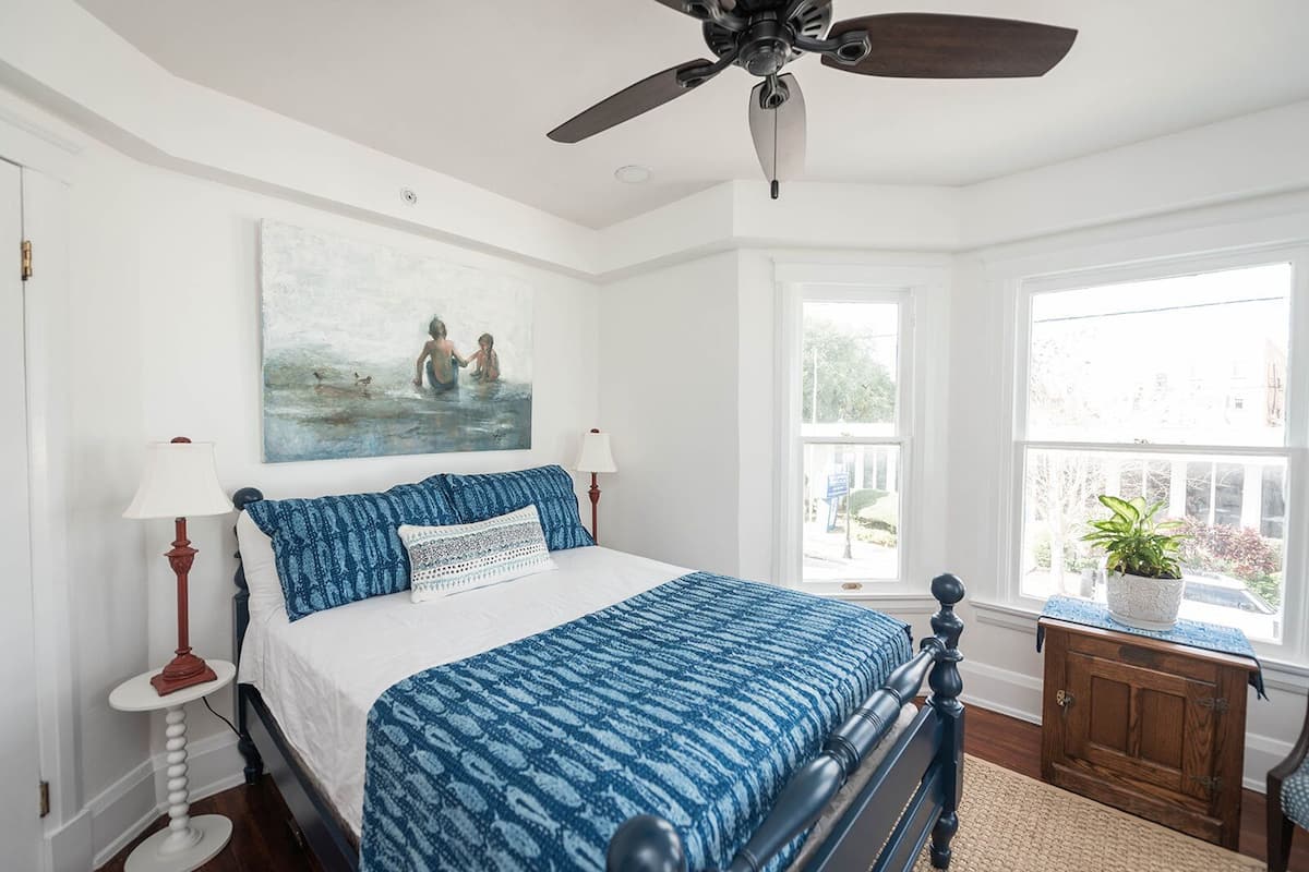 A bedroom with a bed, a ceiling fan, a window with a view, and a painting on the wall.