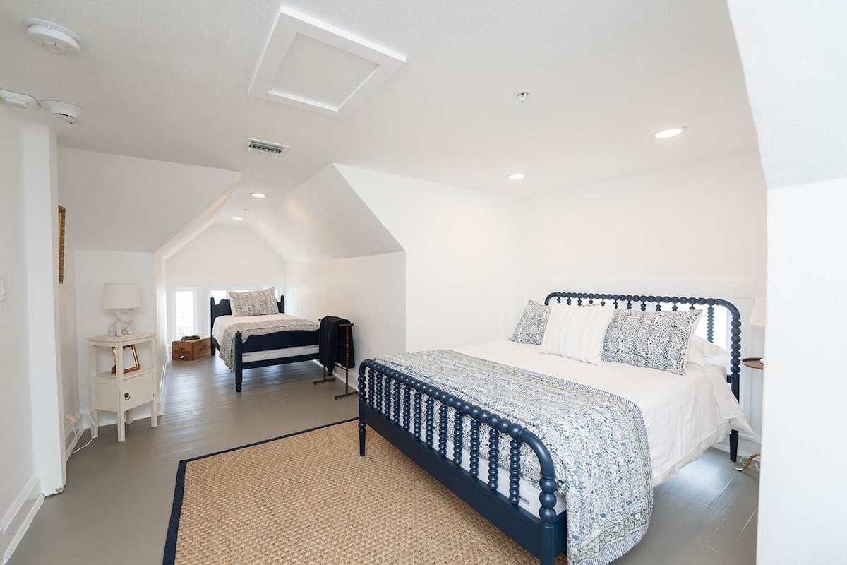 A spacious bedroom with a large bed, a smaller bed in the background, a bedside table with a lamp, and a ceiling light.