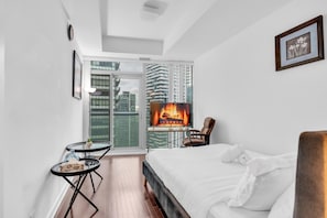 A modern hotel room with a large bed, a small table, a chair, a TV with a fireplace, and a balcony with a city view. - Maple Square Hotel (Toronto)