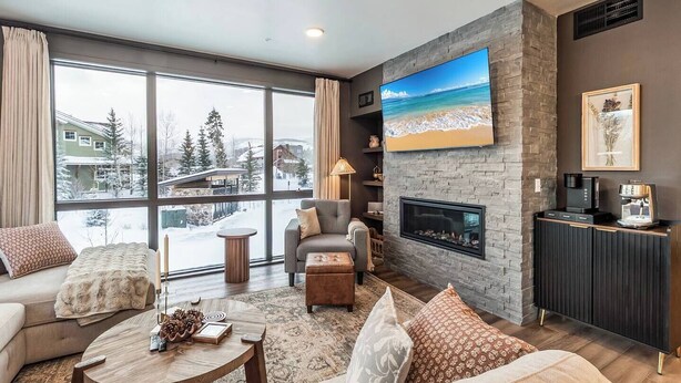 Uma sala de estar moderna com lareira, uma televisão grande e vista para uma paisagem nevada através de amplas janelas.