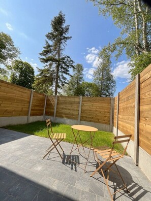 An outdoor patio with a round table and two chairs, surrounded by a wooden fence and greenery. - At Secret des Bois: Air-conditioned house, patio, new appliances. Natural Serenity (Bénifontaine)