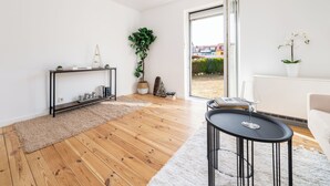 A modern living room with a wooden floor, a black console table, a white sofa, and a large window with a view of buildings and greenery.