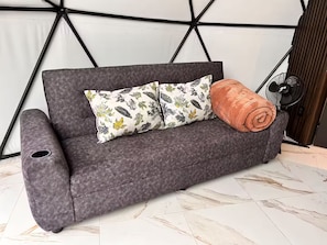 Ein graues Sofa mit einem gemusterten Kissen und einer zusammengerollten Decke, ein Ventilator und ein geometrisches Wanddesign.