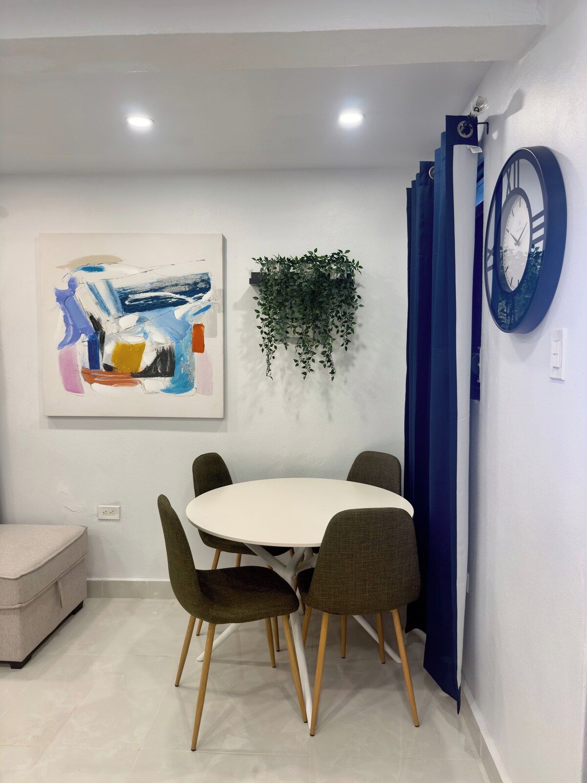 A dining area with a white table and four chairs, a wall-mounted clock, a painting, and a hanging plant.