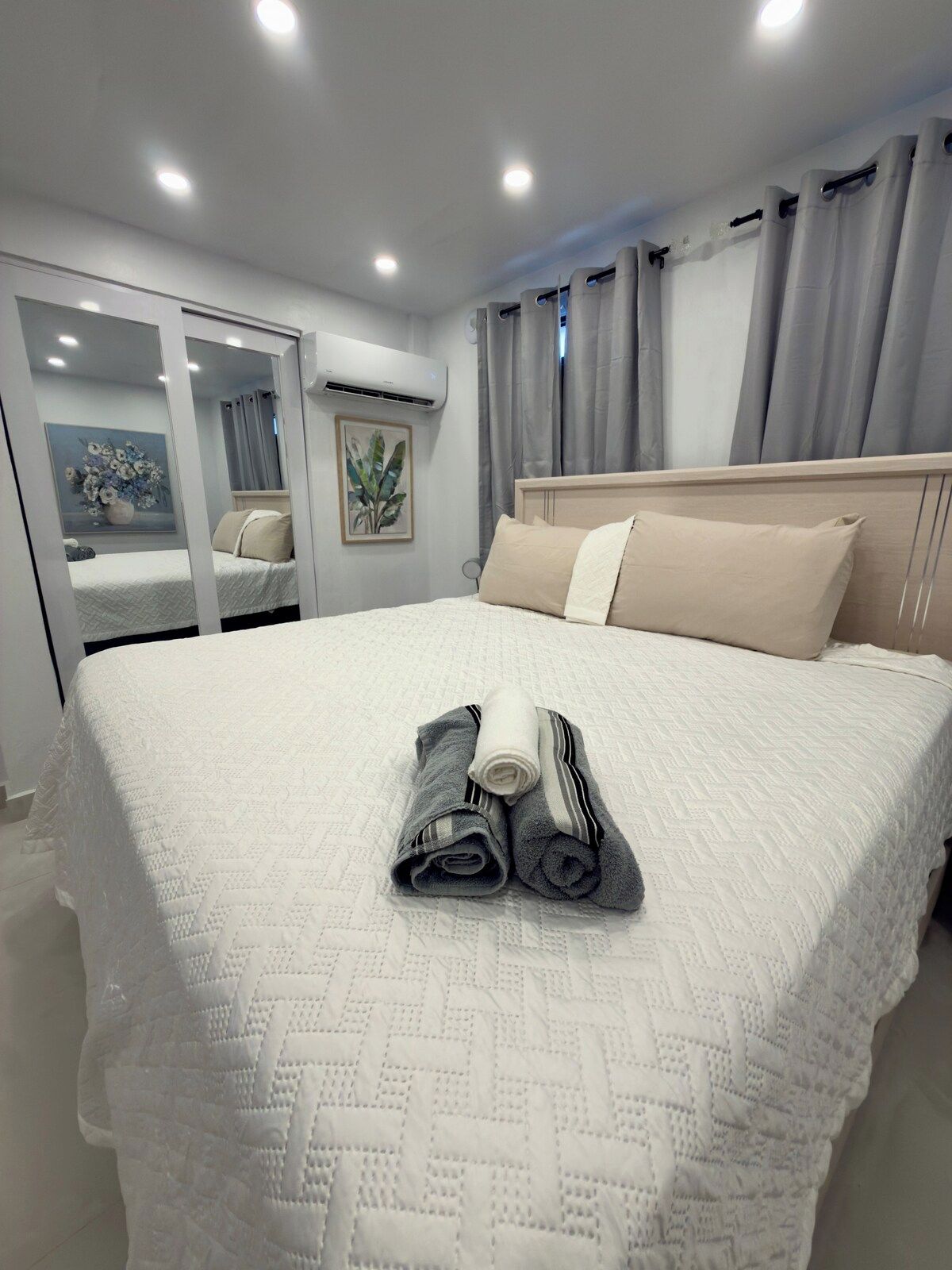 Een opgemaakt bed met opgerolde handdoeken, een nachtkastje en een raam met gordijnen.