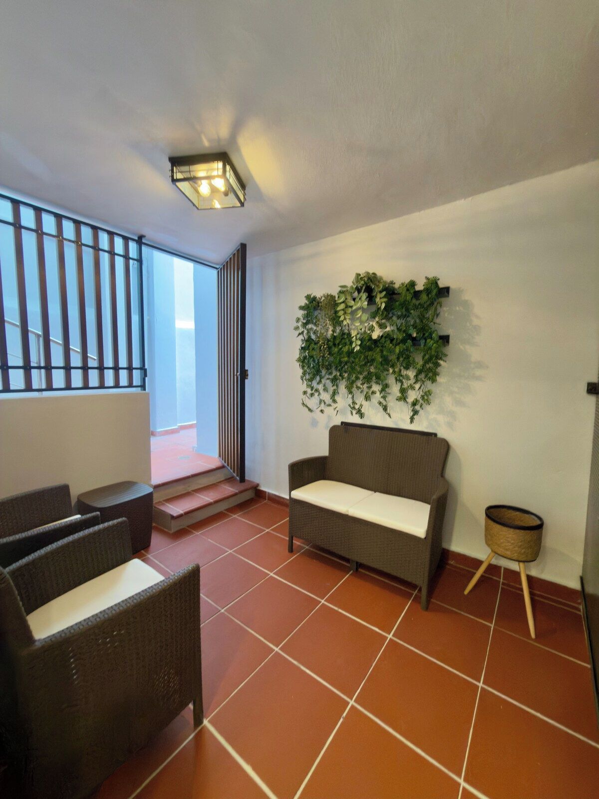 Een kamer met een bank, stoelen, een kleine tafel, een aan de muur bevestigde plant en een trap naar een buitenruimte.