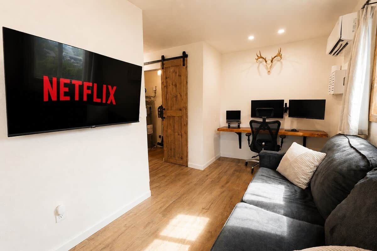 A modern living room with a large flat-screen TV displaying 'NETFLIX,' a wooden door, a desk with two monitors, a leather sofa, and a mounted deer head.