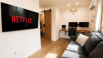 A modern living room with a large flat-screen TV displaying 'NETFLIX,' a wooden door, a desk with two monitors, a leather sofa, and a mounted deer head.