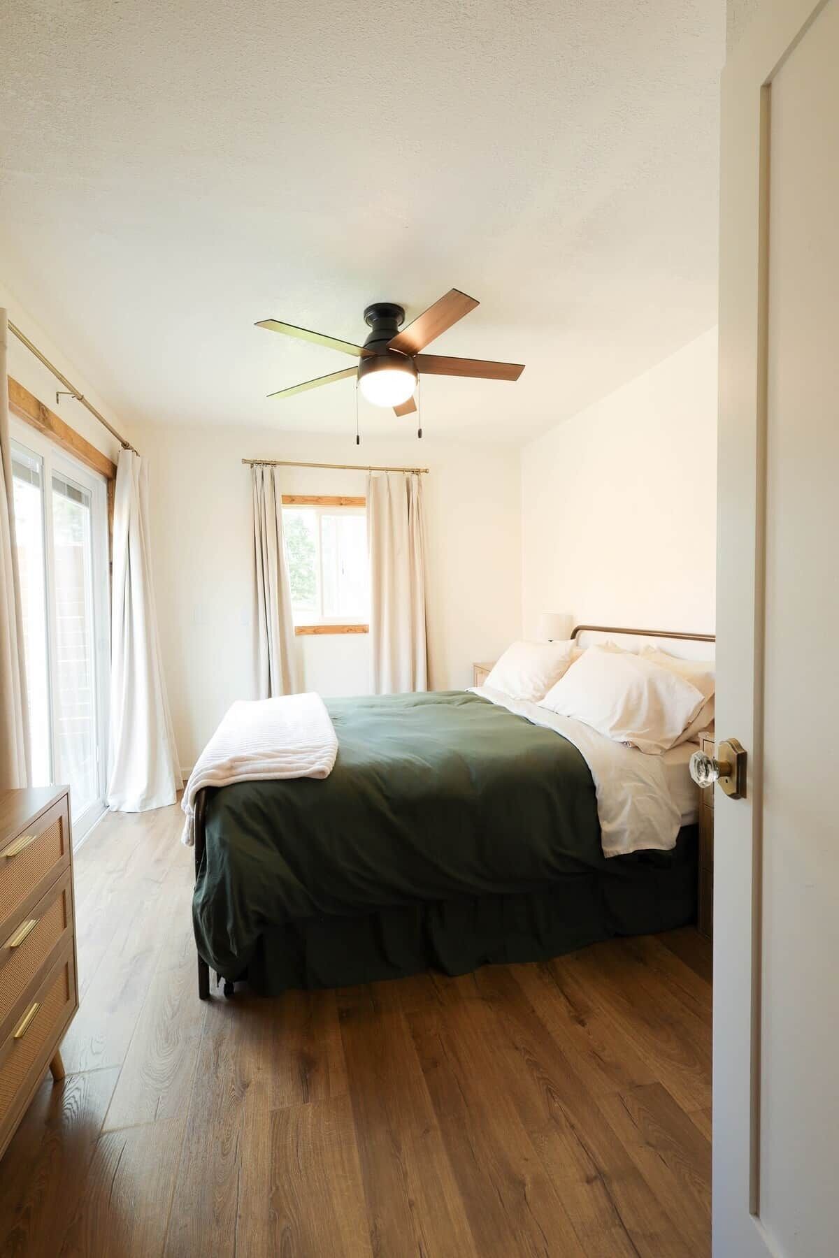 A bedroom with a bed, a ceiling fan, a window with curtains, and a wooden dresser.