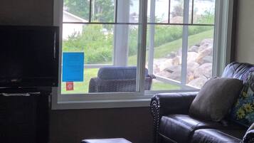 A living room with a television, a sofa, a coffee table, and a window with a view of greenery and a building.