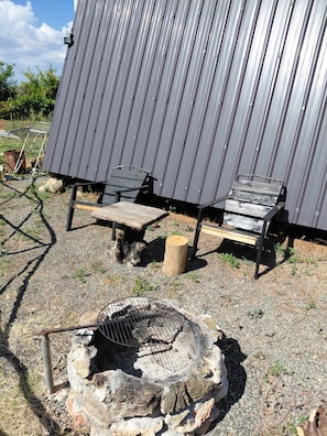 Un edificio de metal con suelo de grava, dos sillas de madera y una parrilla metálica.