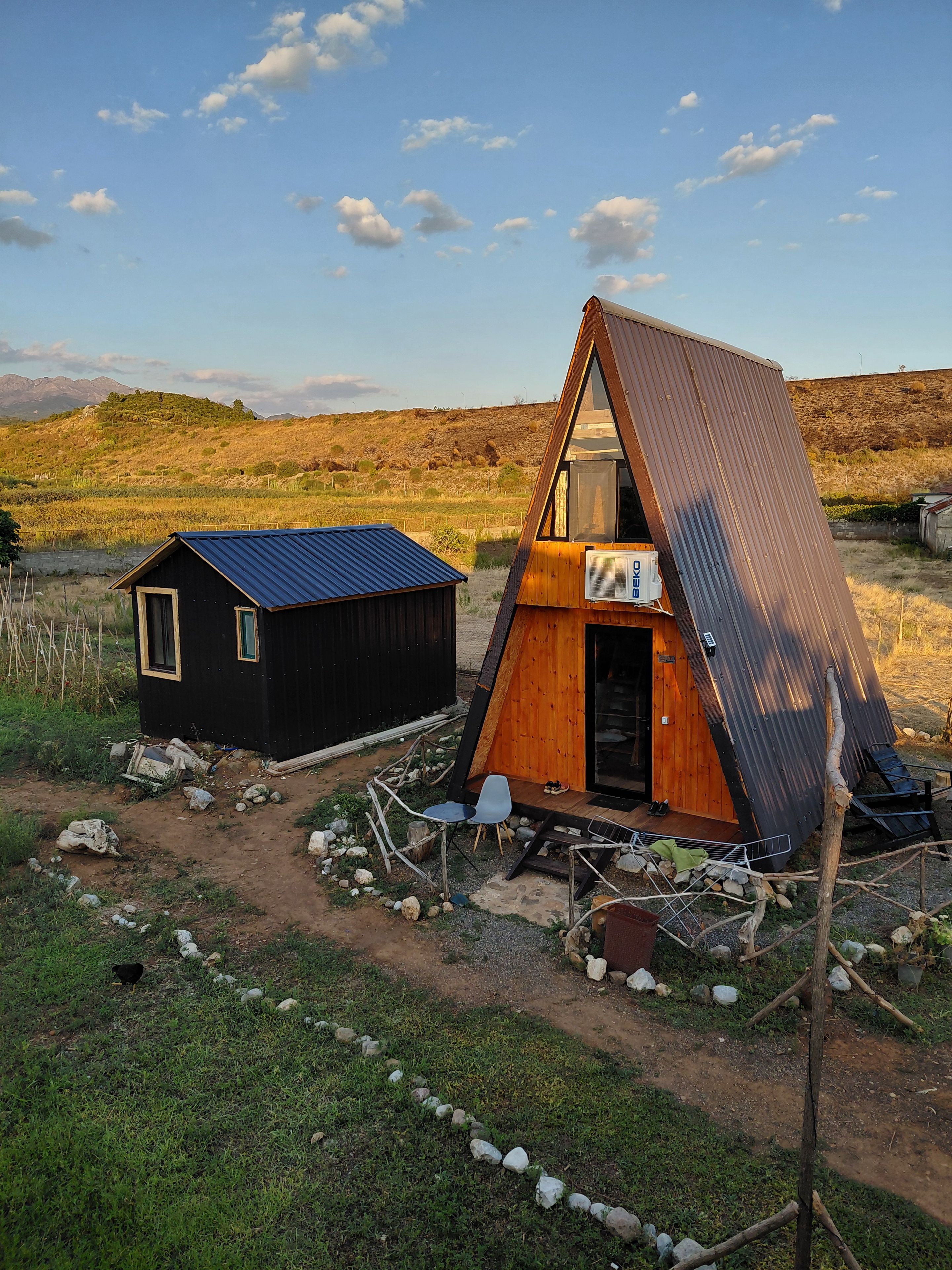 Eine kleine, zweistöckige Holzhütte mit einem geneigten Dach, einem separaten Gartenhaus und einem Feldweg.