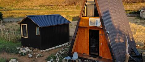 A small, two-story cabin with a sloped roof, a separate shed, and a dirt path.