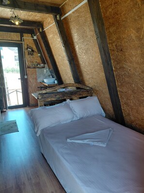 A room with a bed, wooden beams, a wooden table with a sink, and a view of the outside.