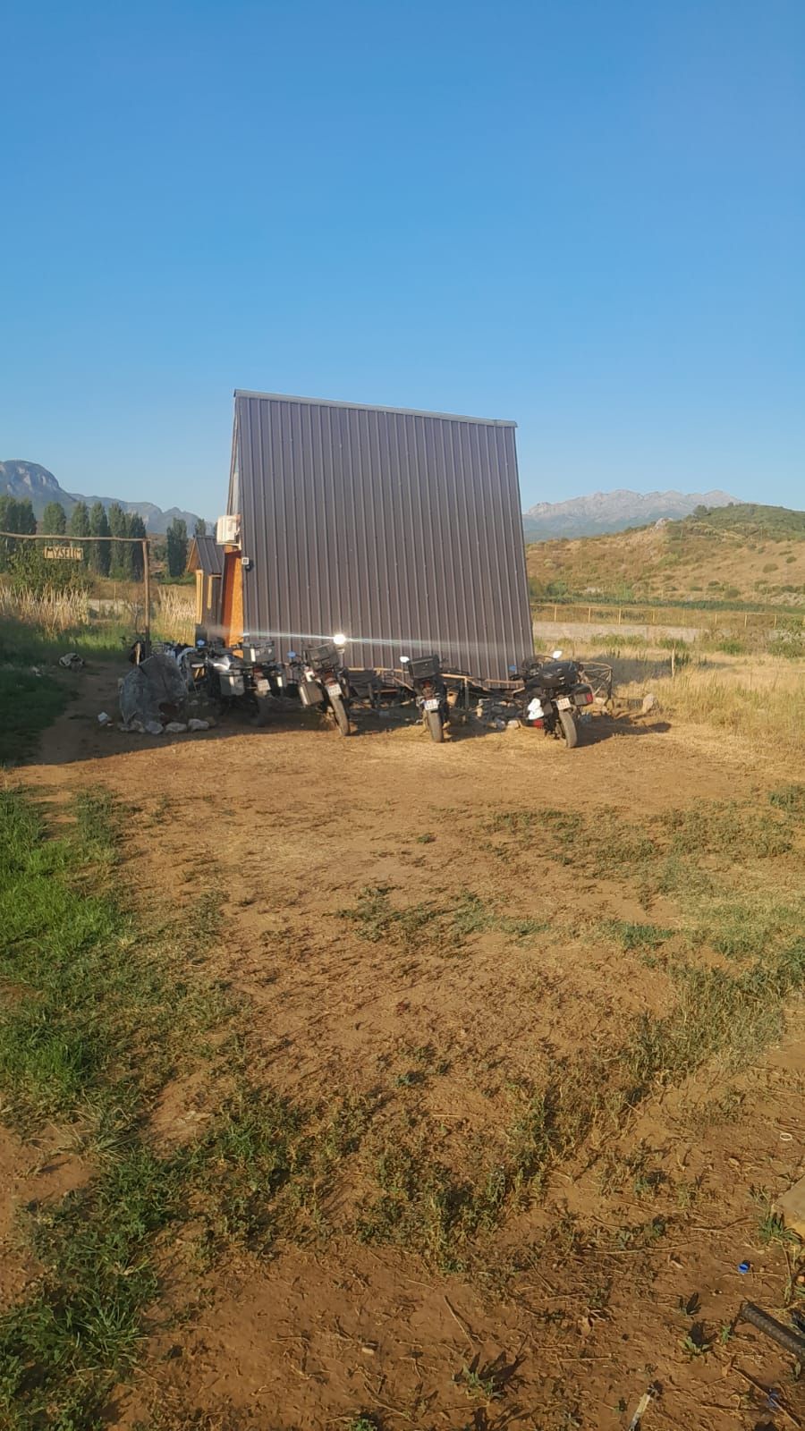 Una estructura metálica grande y rectangular con techo plano, posiblemente un edificio de almacenamiento o servicios, ubicada en un campo seco y cubierto de hierba con cielo despejado y montañas distantes al fondo.