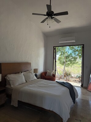 Une chambre  à coucher comprenant un lit, une table de chevet, une chaise, un ventilateur de plafond et une fenêtre donnant sur un paysage verdoyant.